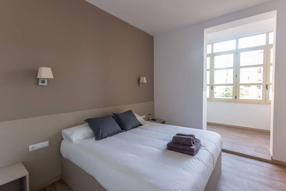 Een hotelkamer met een bed, twee bedlampen, een nachtkastje en een raam met uitzicht.