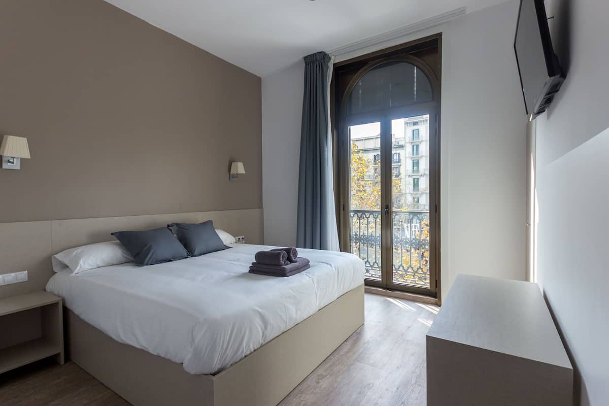 Een moderne hotelkamer met een groot bed, een nachtkastje, een stoel en een balkon met uitzicht op gebouwen en bomen.