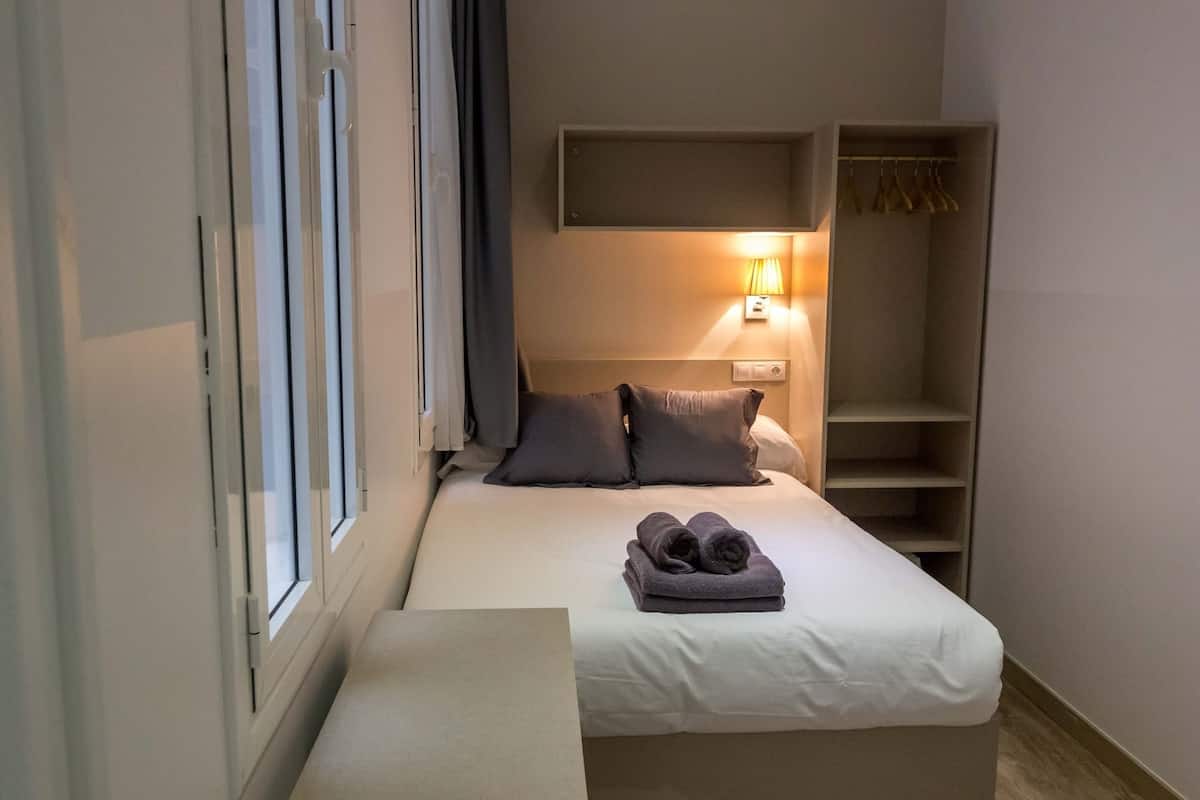 Een kleine, netjes ingerichte slaapkamer met een bed, een bedlampje, een plankje en een raam.