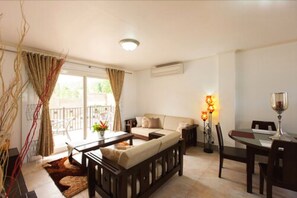 A living room with a sofa, coffee table, and dining table. There is a balcony with curtains, a lamp, and a window. - Praslin Holiday Home (Praslin Island)