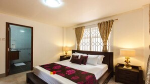 A hotel room with a large bed, a wooden headboard, two bedside tables with lamps, a window with curtains, and a bathroom visible through an open door. - Praslin Holiday Home (Praslin Island)
