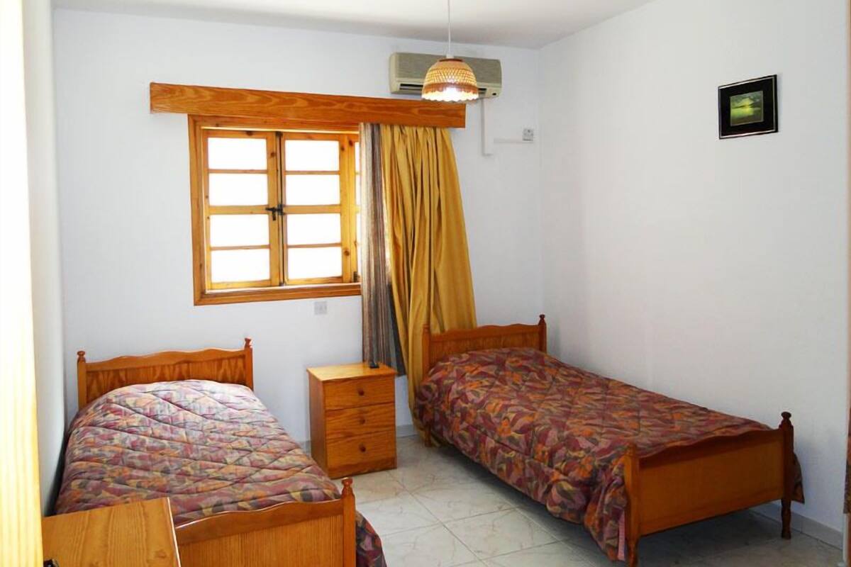 Un dormitorio pequeño y bien iluminado con dos camas de madera, una ventana con cortinas, una mesita de noche y una lámpara colgante.