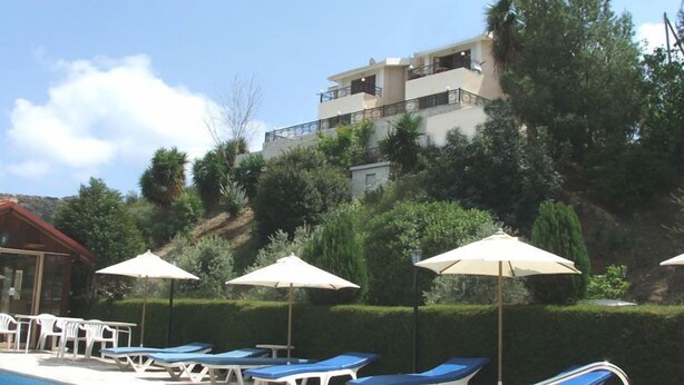 A pool area with lounge chairs and umbrellas, a building with balconies, and a hillside with trees. - Ampelokipi Holiday Apartments (Pissouri)