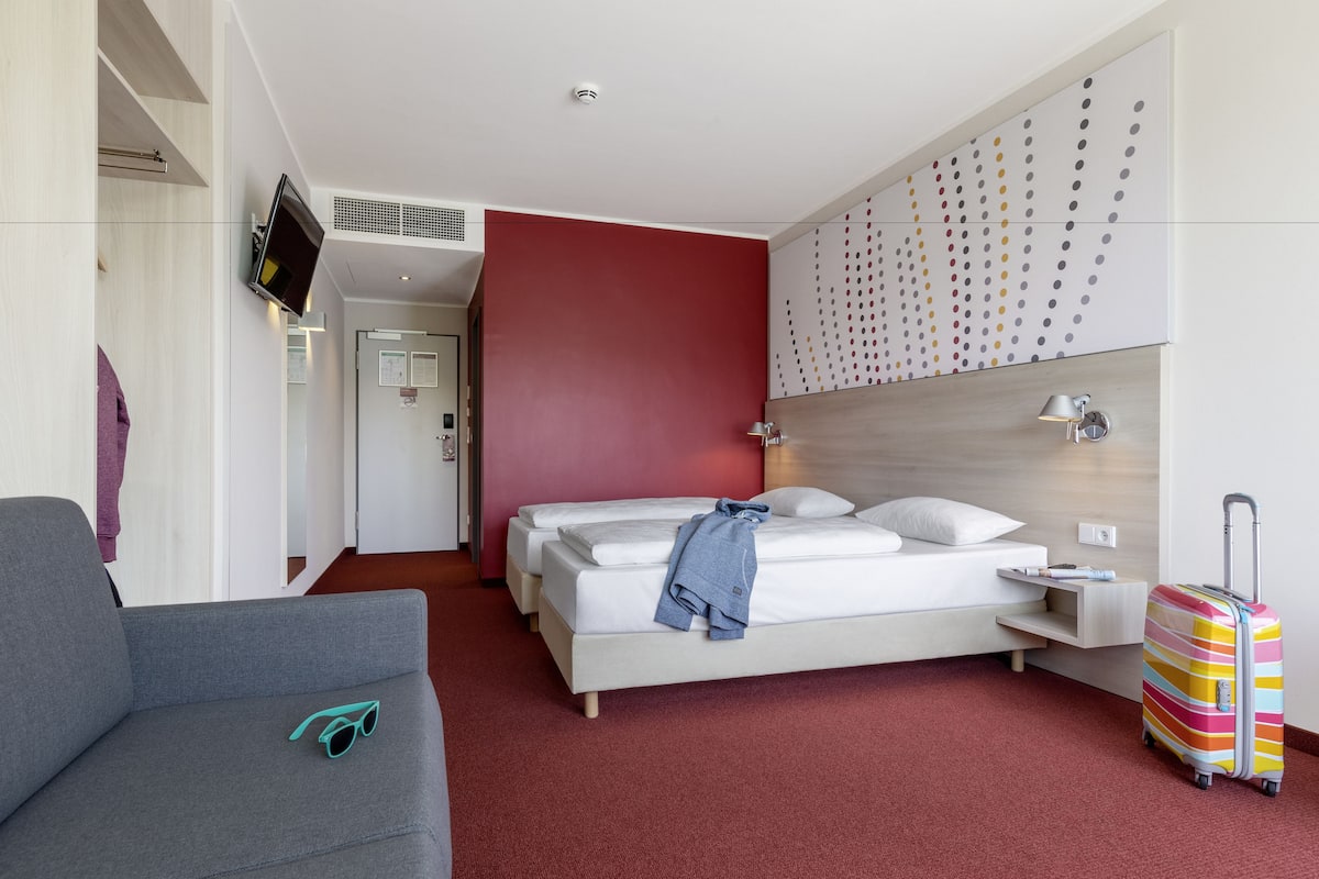 Quarto de hotel com uma cama, um sofá cinzento, mala colorida e televisão na parede.