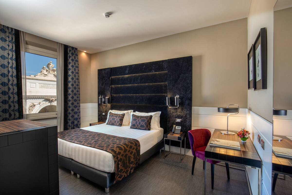 Une chambre d’hôtel moderne avec un grand lit, un bureau et une chaise. Une fenêtre offre une vue sur une statue située à l’extérieur.