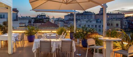An outdoor dining area with tables and chairs, potted plants, and a view of the cityscape.