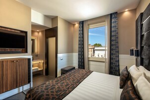 A hotel room with a bed, a TV mounted on the wall, a window with curtains, and a view of the city.