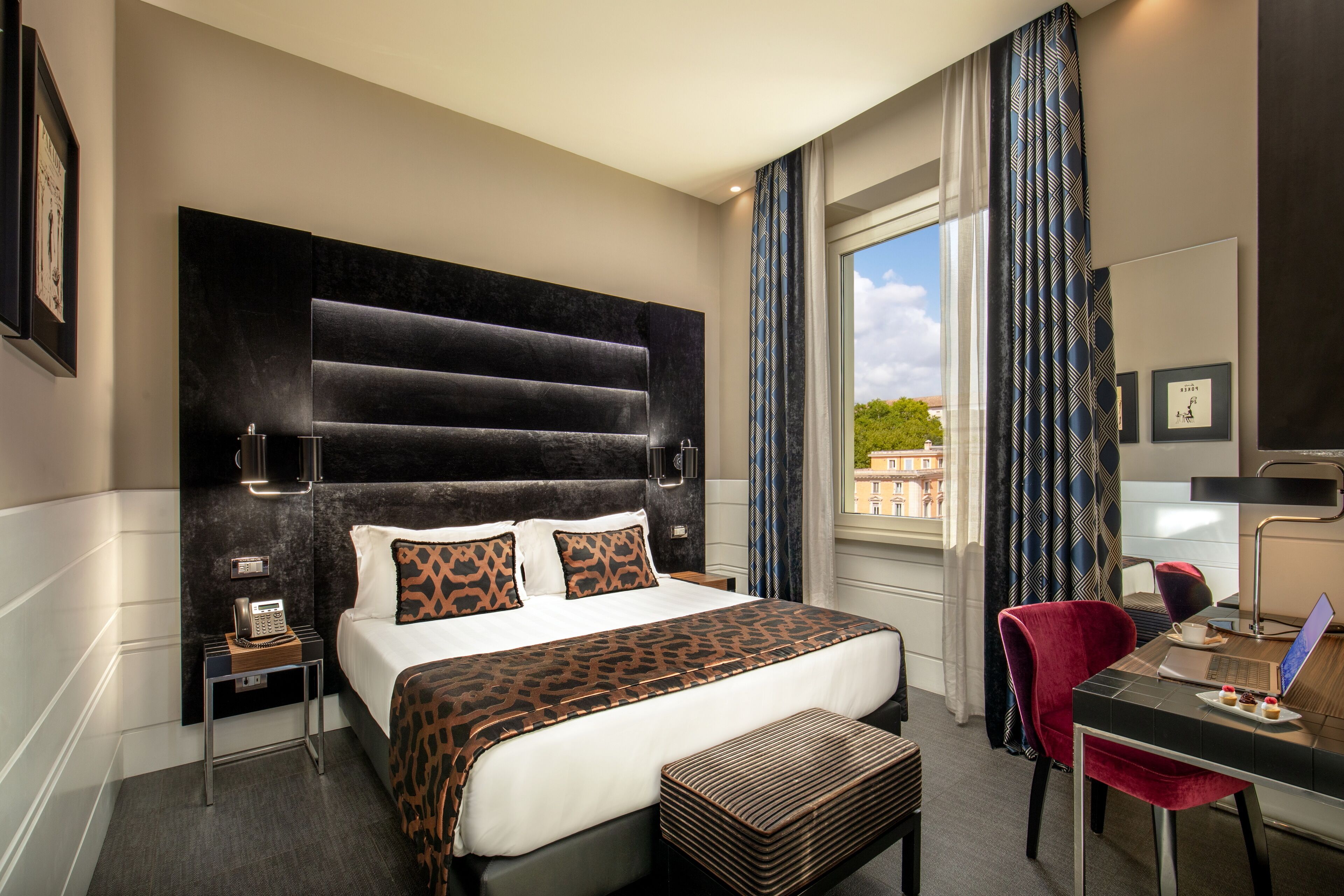 Quarto de hotel com uma cama grande, uma escrivaninha com um laptop, uma cadeira, um telefone e uma janela com cortinas.