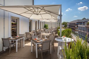 An outdoor dining area with multiple tables and chairs, large umbrellas, and a view of the cityscape.