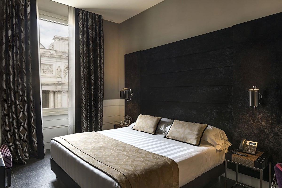 Une chambre d’hôtel avec un grand lit, un mur d’accent de couleur sombre, une vue sur un immeuble par la fenêtre et un téléphone sur la table de chevet.