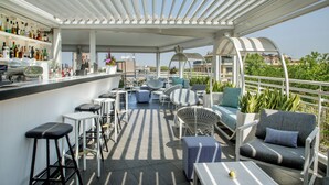 A rooftop bar with white bar stools, seating areas with cushions, and a variety of plants. - The Tribune, part of JdV by Hyatt (Rome)