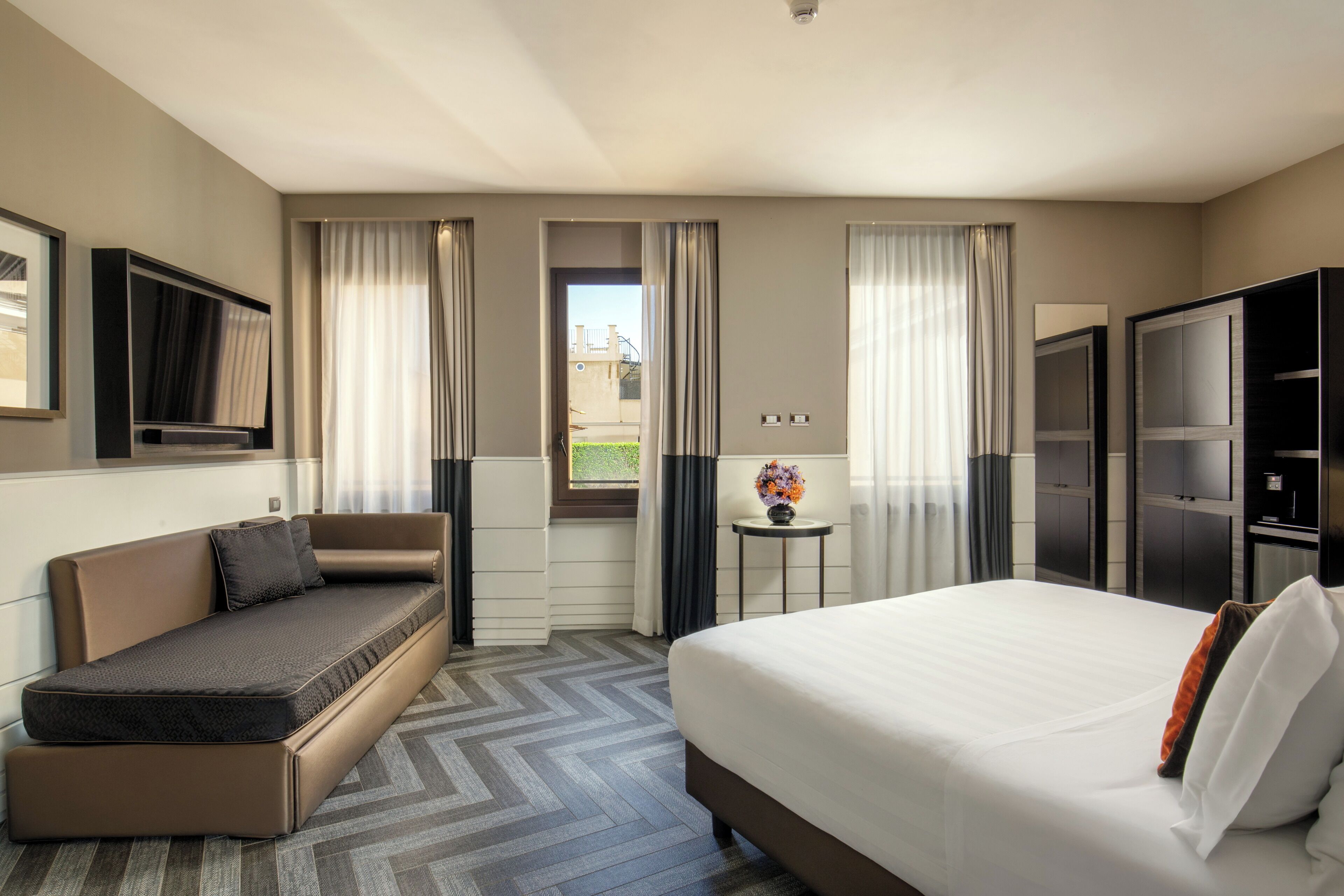 Une chambre d’hôtel avec un lit, un canapé, une télévision et une petite table sur laquelle est posé un vase de fleurs.