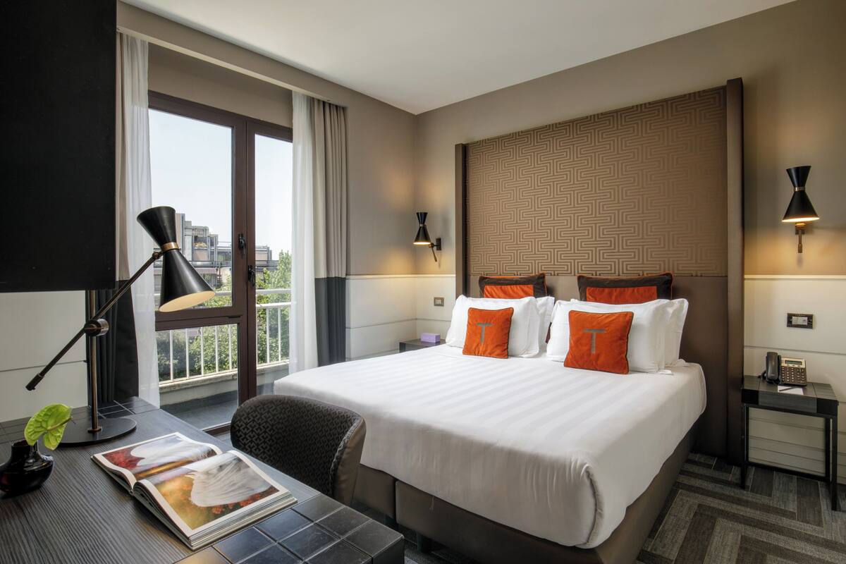 Une chambre d’hôtel avec un grand lit, deux oreillers, une table de chevet avec une lampe, une chaise, un bureau avec un magazine et une vue sur la ville par la fenêtre.