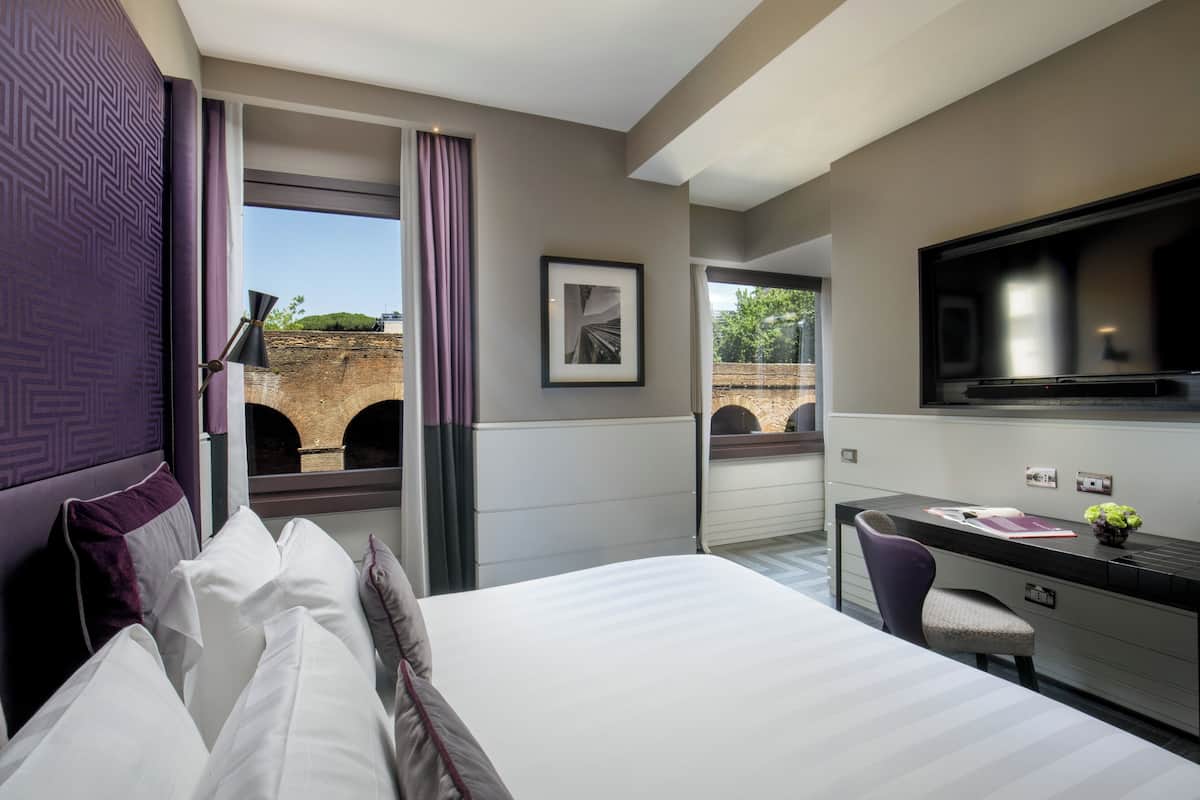 Une chambre d’hôtel avec un lit, un bureau et une télévision. Il y a une fenêtre avec des rideaux, et de cette fenêtre, on peut voir un mur de briques et de la verdure à l’extérieur.
