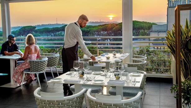 A restaurant with outdoor seating overlooking a scenic view, tables set with white tableware, and a waiter attending to guests. - The Tribune, part of JdV by Hyatt (Rome)