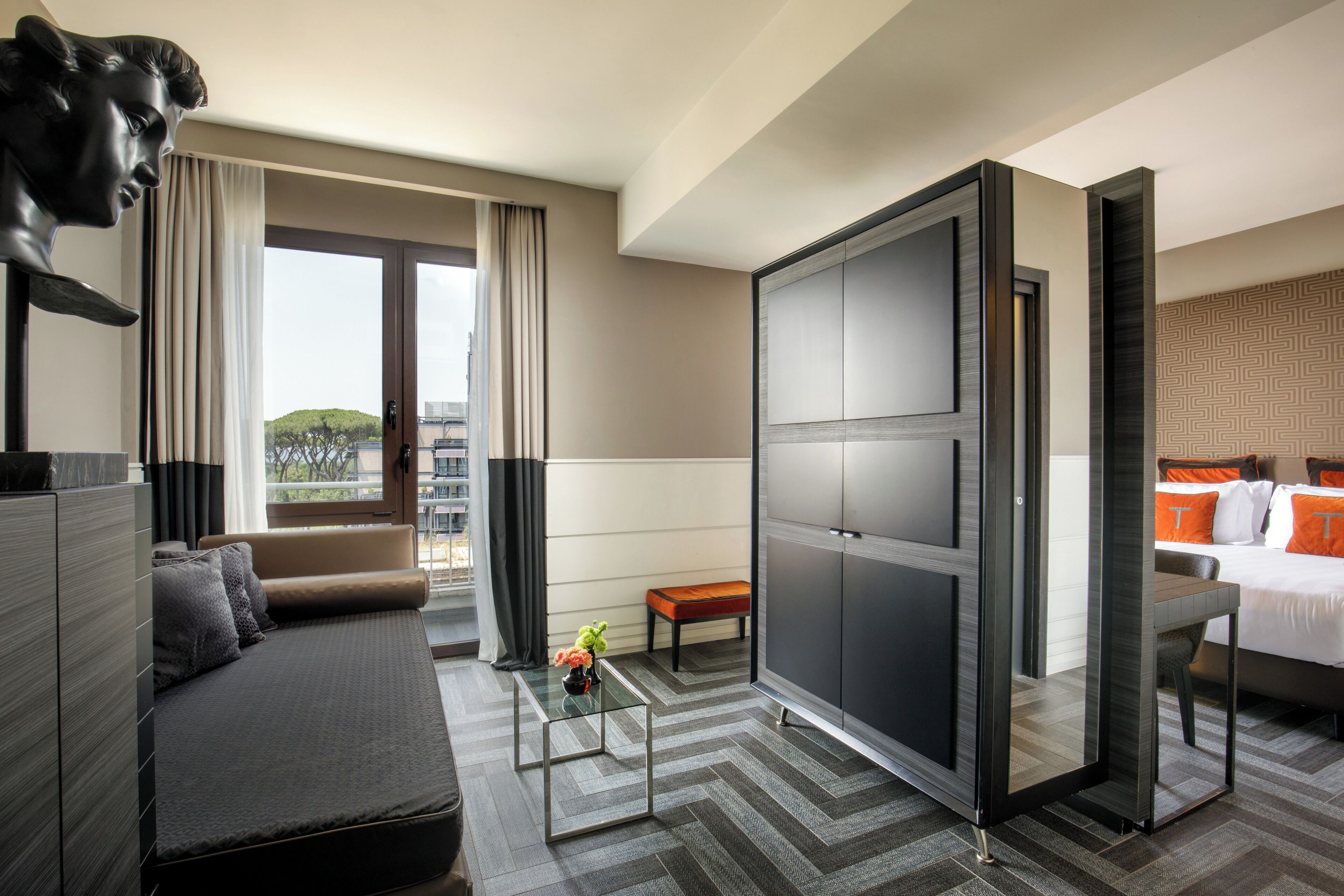 A modern hotel room with a sofa, a glass coffee table, a bed with orange pillows, and a large wardrobe.