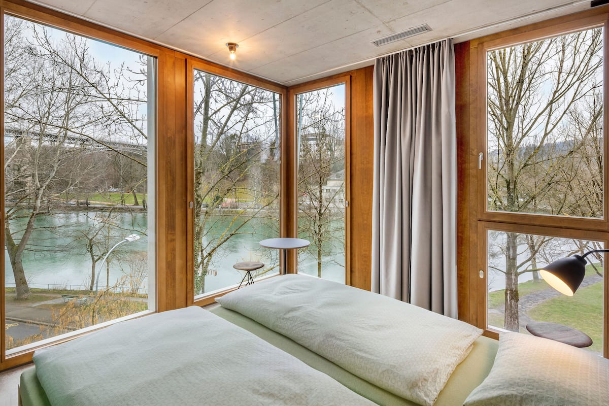 A room with large windows, a bed, a table, and a lamp.