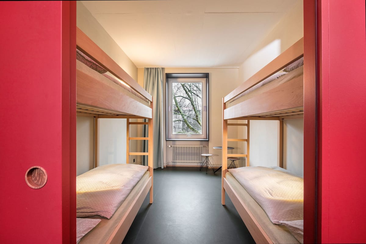 A dormitory room with bunk beds, a small table, and a window with a view of trees.