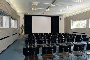 Un salón de conferencias con filas de sillas, una pantalla grande y ventanas.