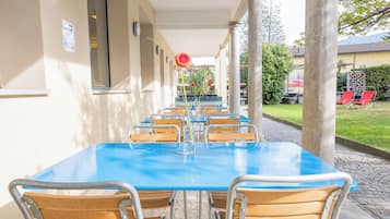 Un área de comedor al aire libre cubierta con una mesa azul y sillas de madera, rodeada de columnas y vistas a un edificio y follaje.