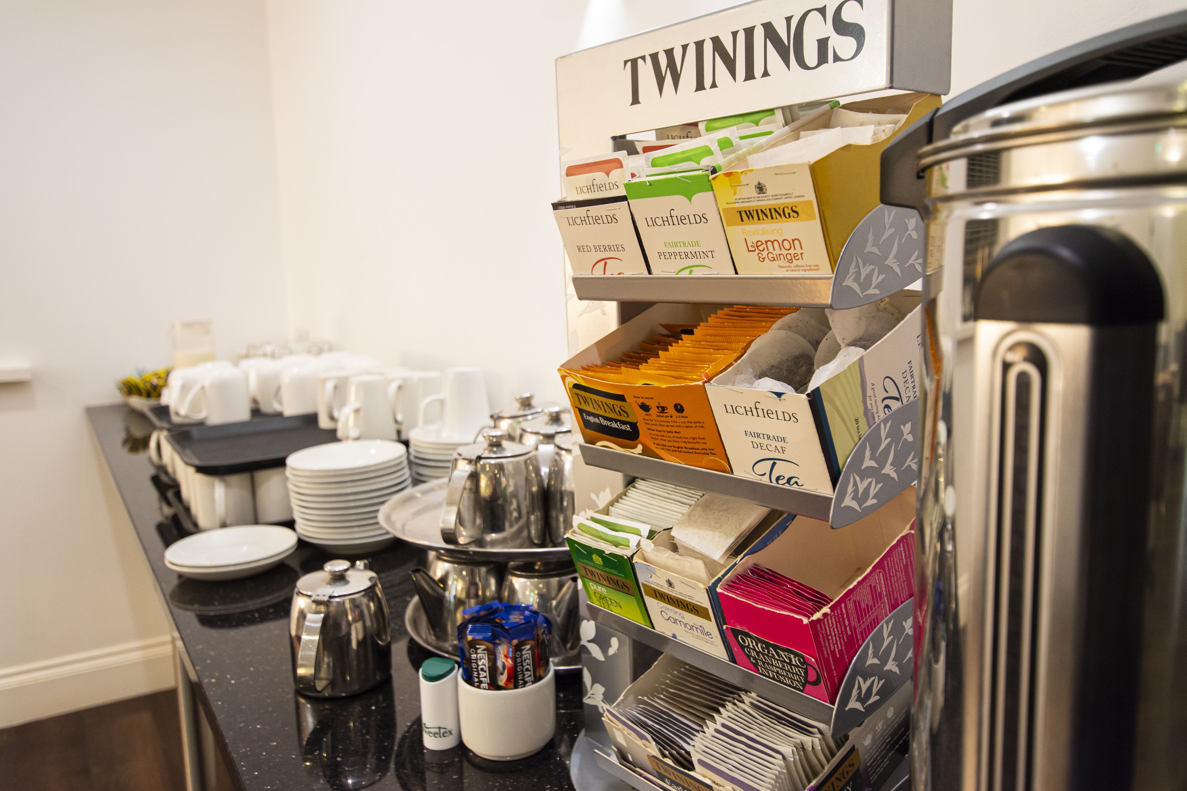 Montaje tipo bufé con diversas opciones de comida y bebida, incluyendo una máquina de café y un cartel que dice "TWININGS".