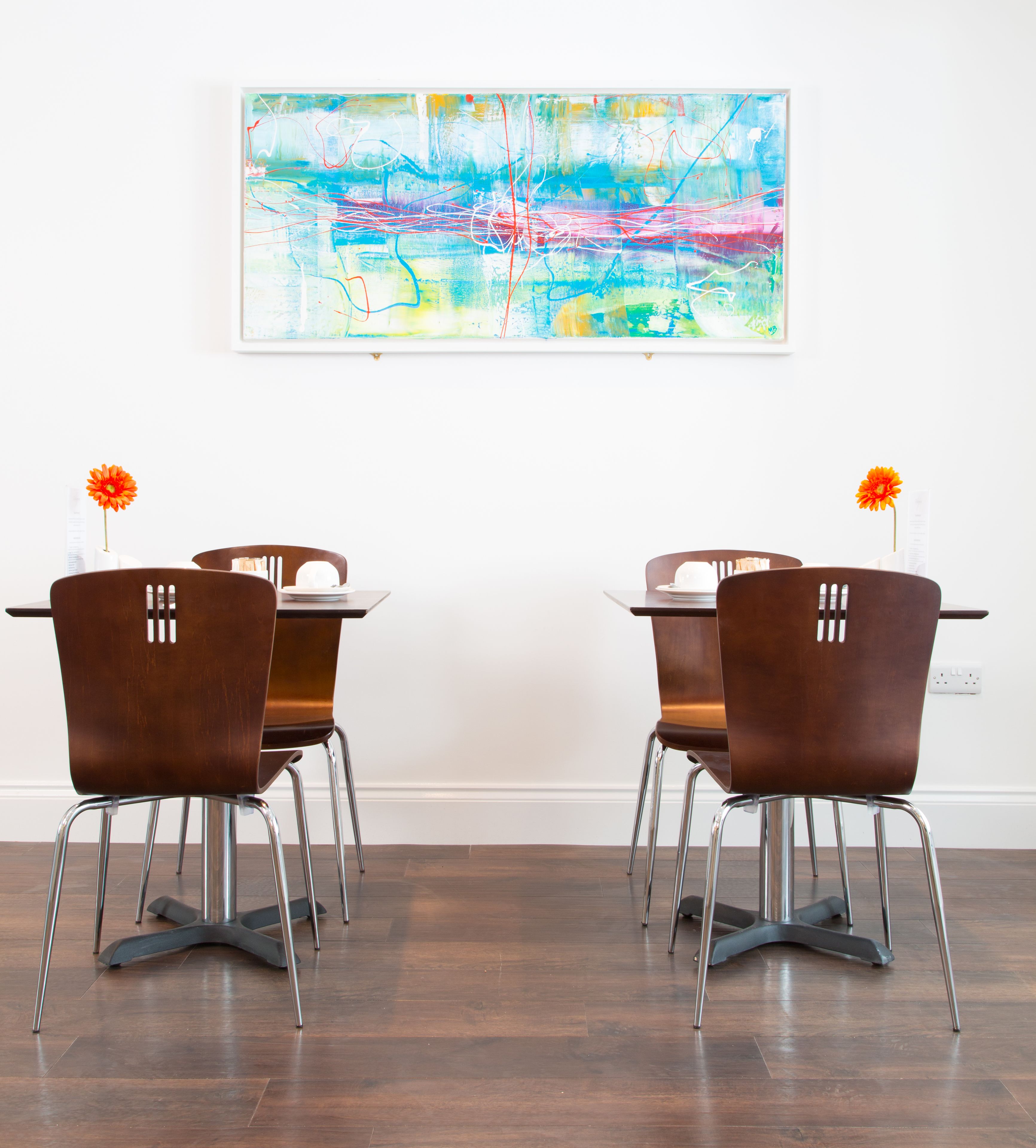Un coin repas avec quatre chaises marron et une table, un tableau abstrait aux couleurs vives accroché au mur, et un vase à fleurs posé sur la table.