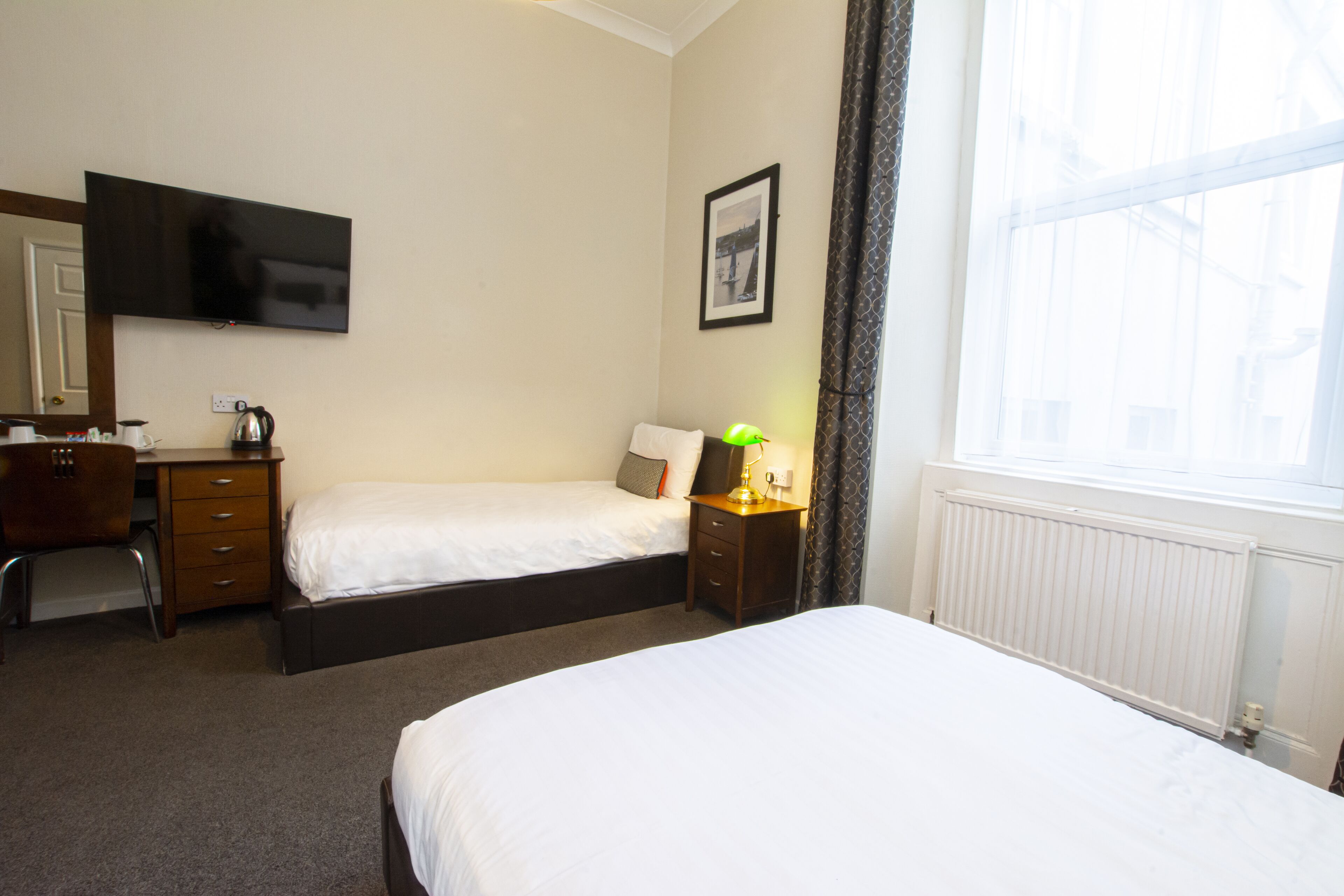 A hotel room with two single beds, a desk with a chair, a television mounted on the wall, a framed picture, and a window with curtains.