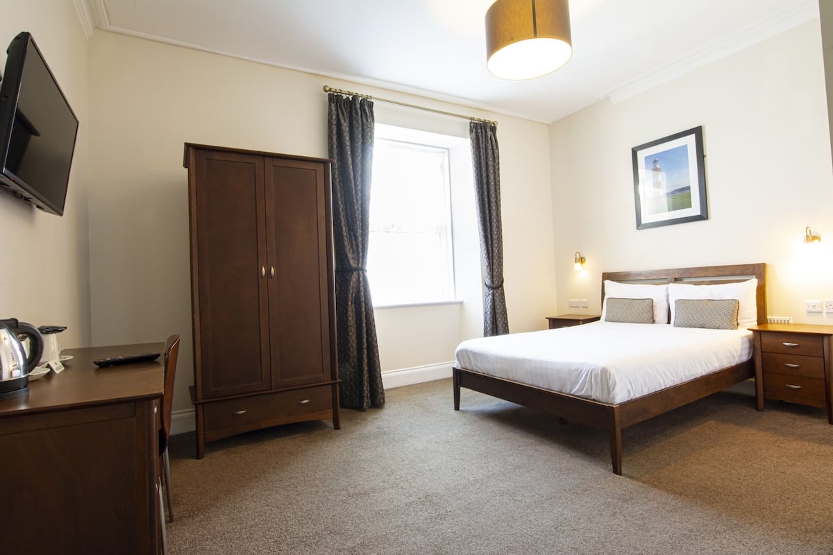 A hotel room with a bed, a wardrobe, a desk with a kettle, a television, and a window with curtains.