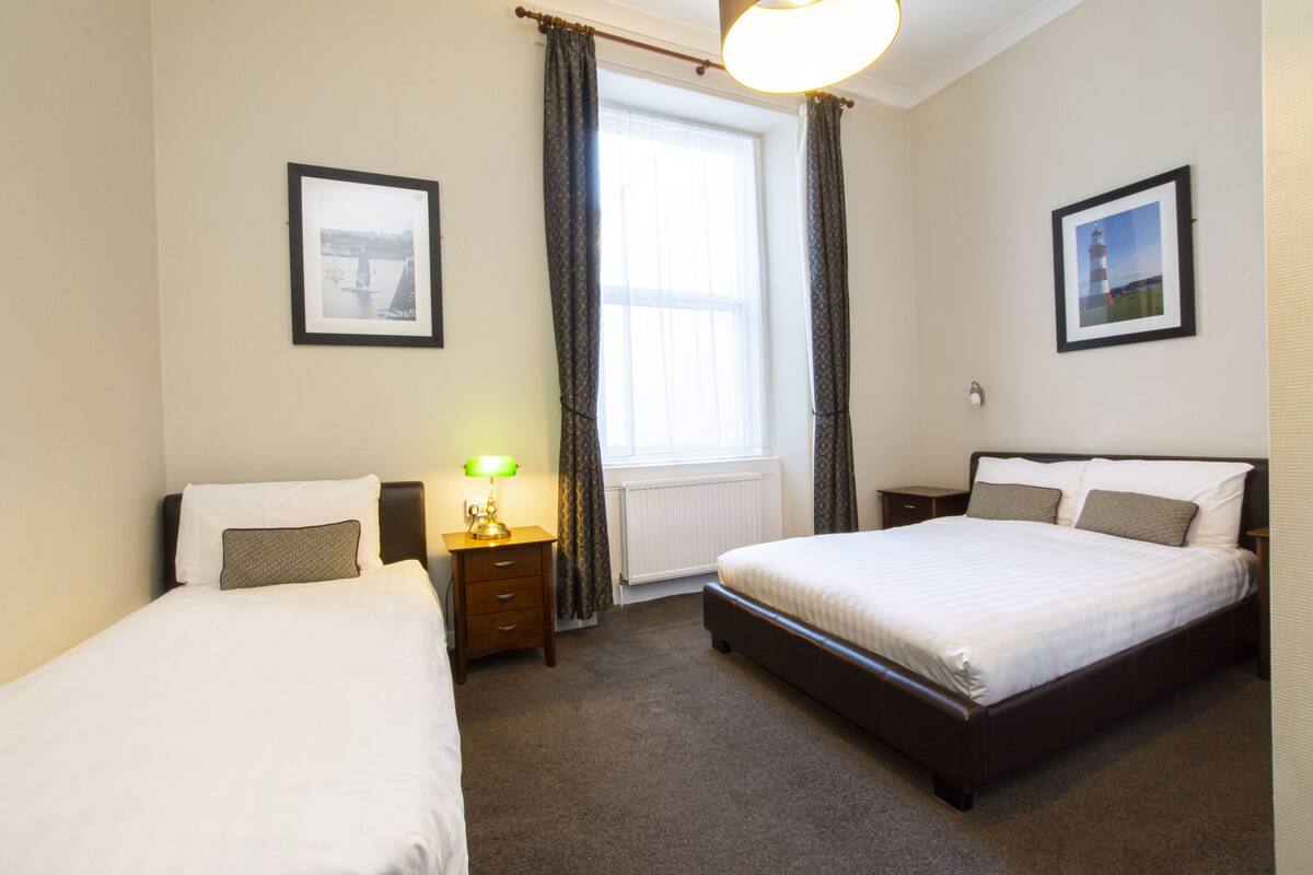 A hotel room with two single beds, a nightstand with a lamp, a window with curtains, and framed pictures on the walls.