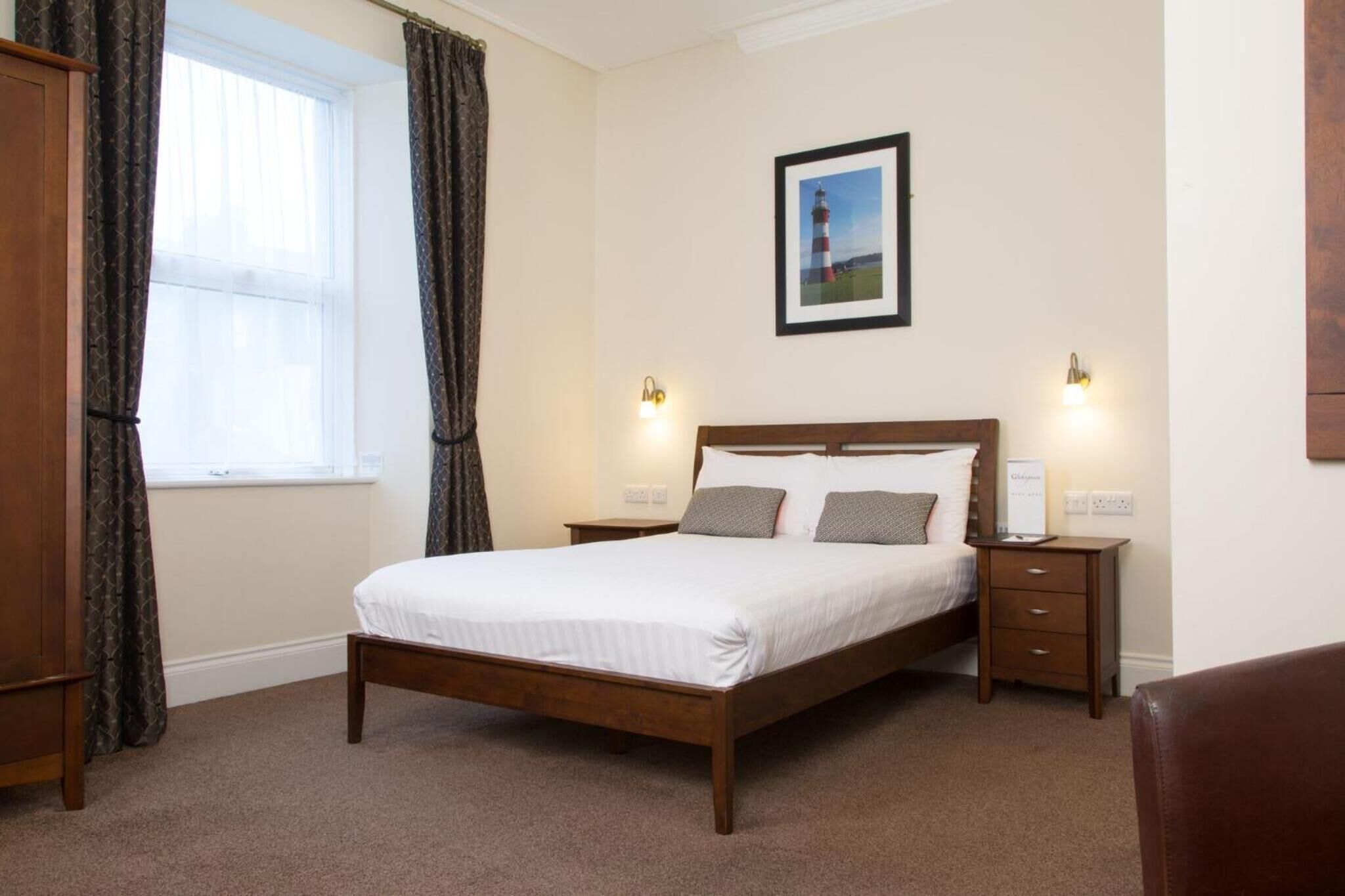 A neatly arranged bedroom with a wooden bed, two bedside tables with lamps, a framed picture on the wall, and a window with curtains.