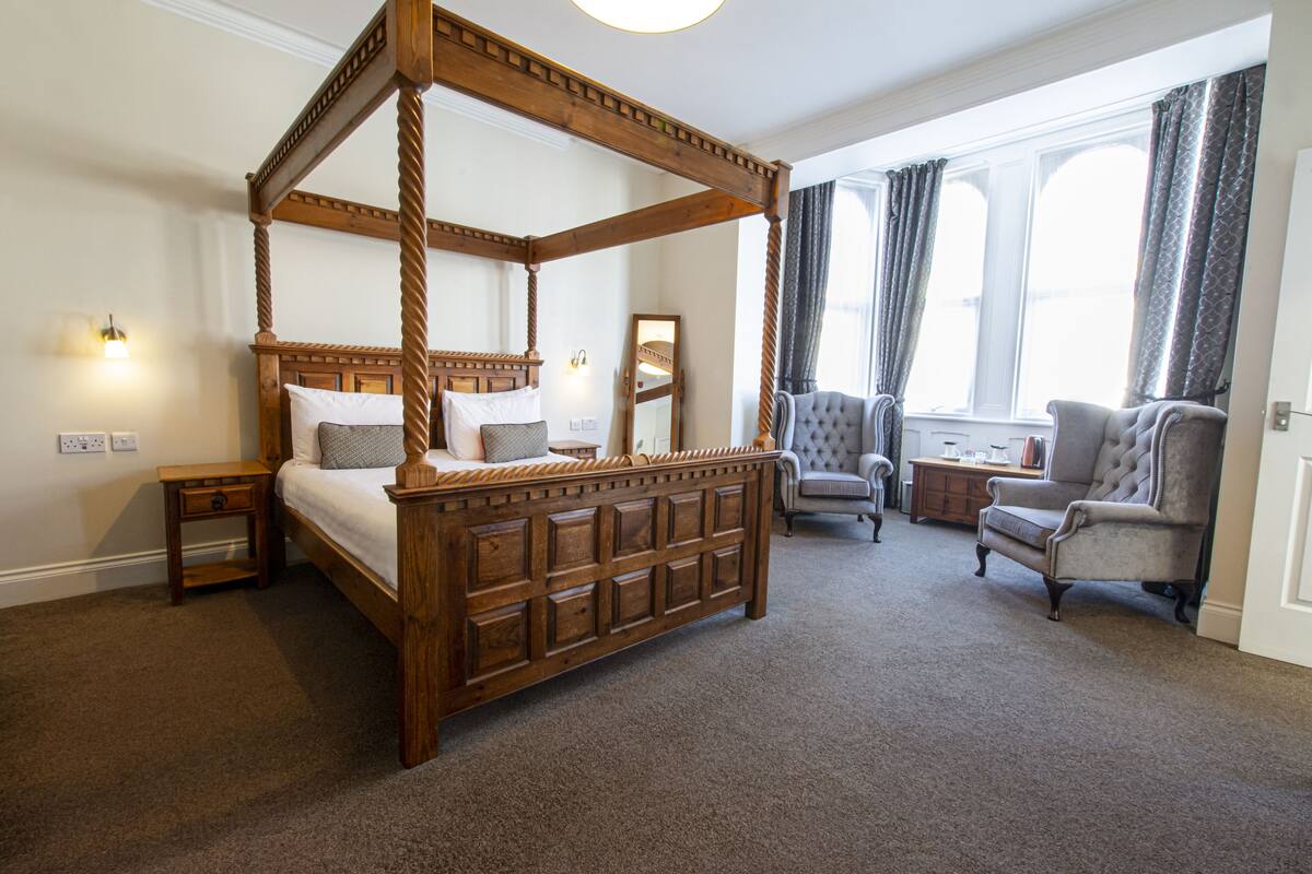 A four-poster bed with a wooden frame, a bedside table, a chair, and a small table with a lamp.