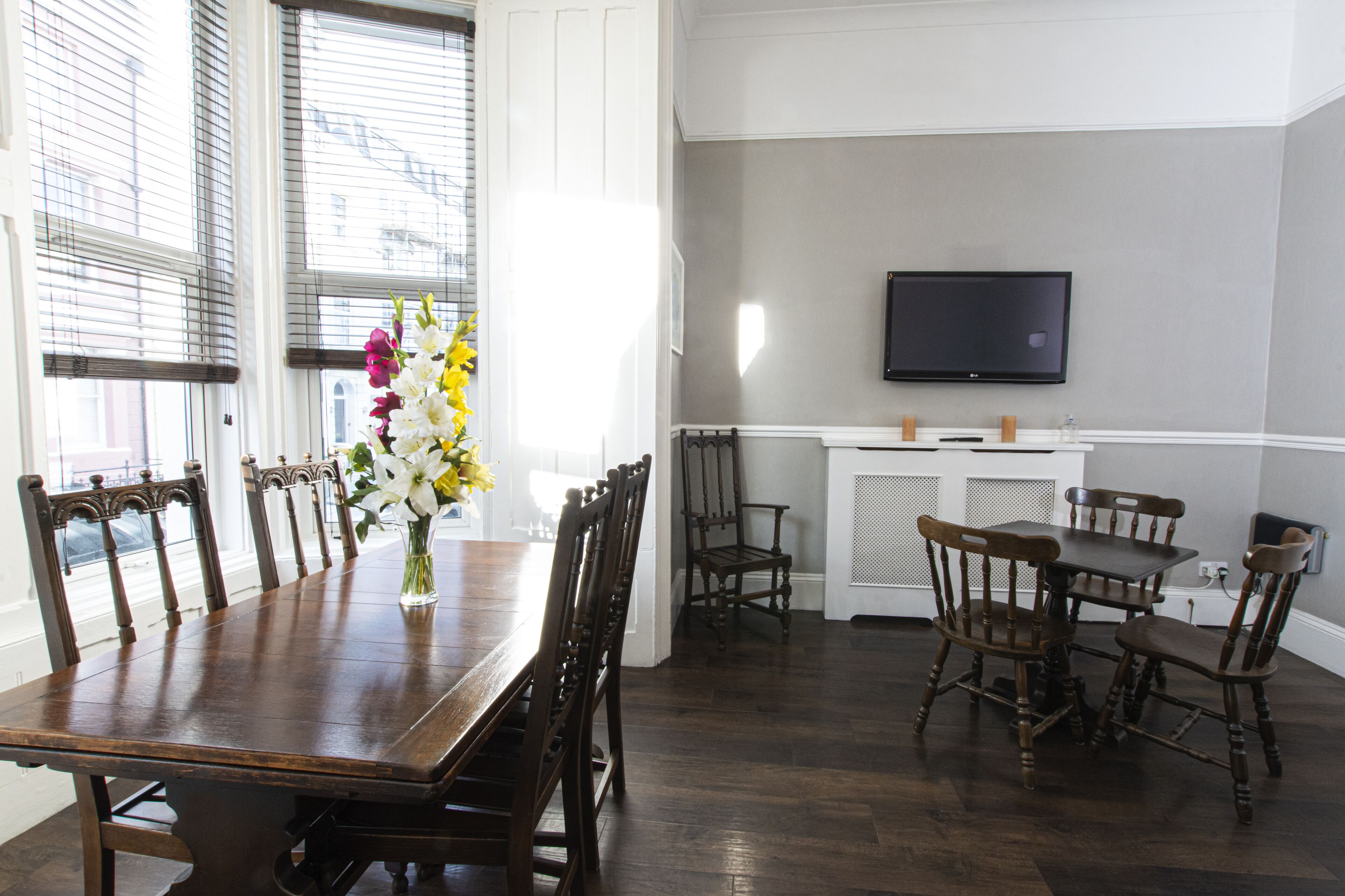 A dining area with a wooden table and chairs, a vase of flowers, and a television mounted on the wall.