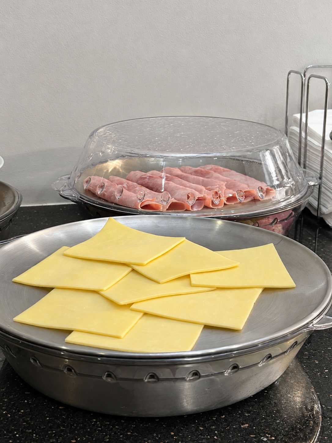 A kitchen counter with a metal pan containing sliced yellow cheese and a glass container with sliced ham.