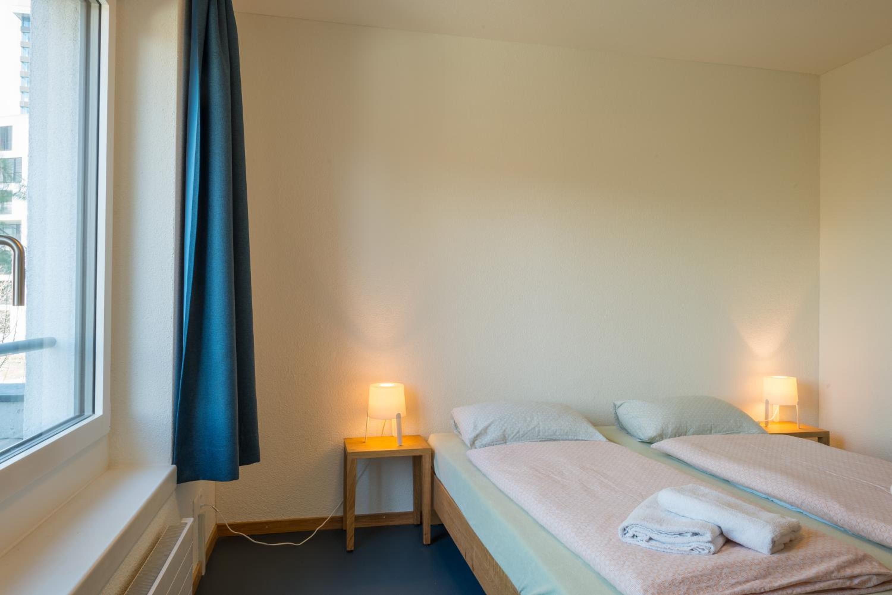 Habitación de hotel con una cama, dos mesitas de noche con lámparas, una ventana con cortina y vistas al exterior.