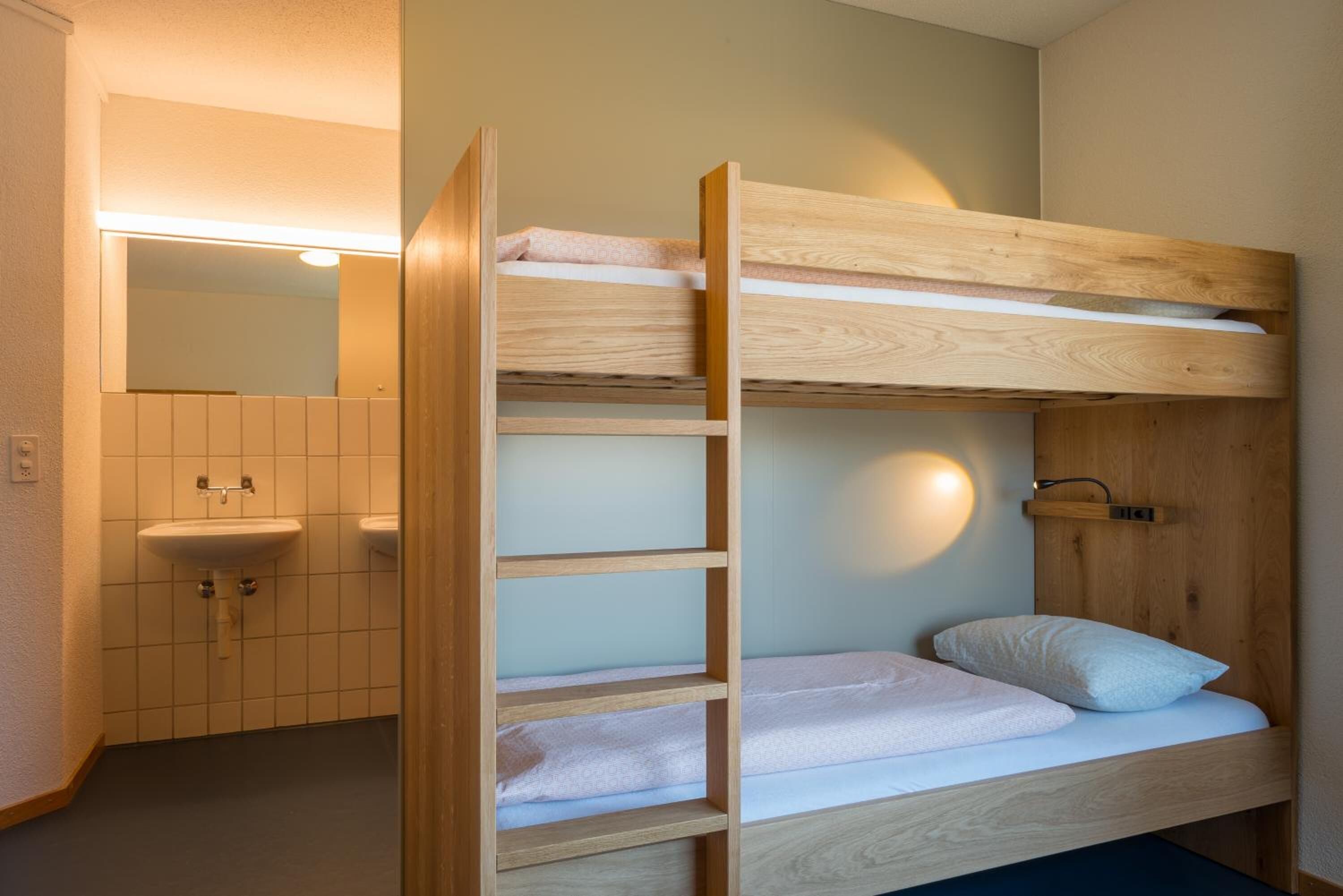 Una cama litera con estructura de madera, un estante pequeño y una mesita de noche con lámpara.