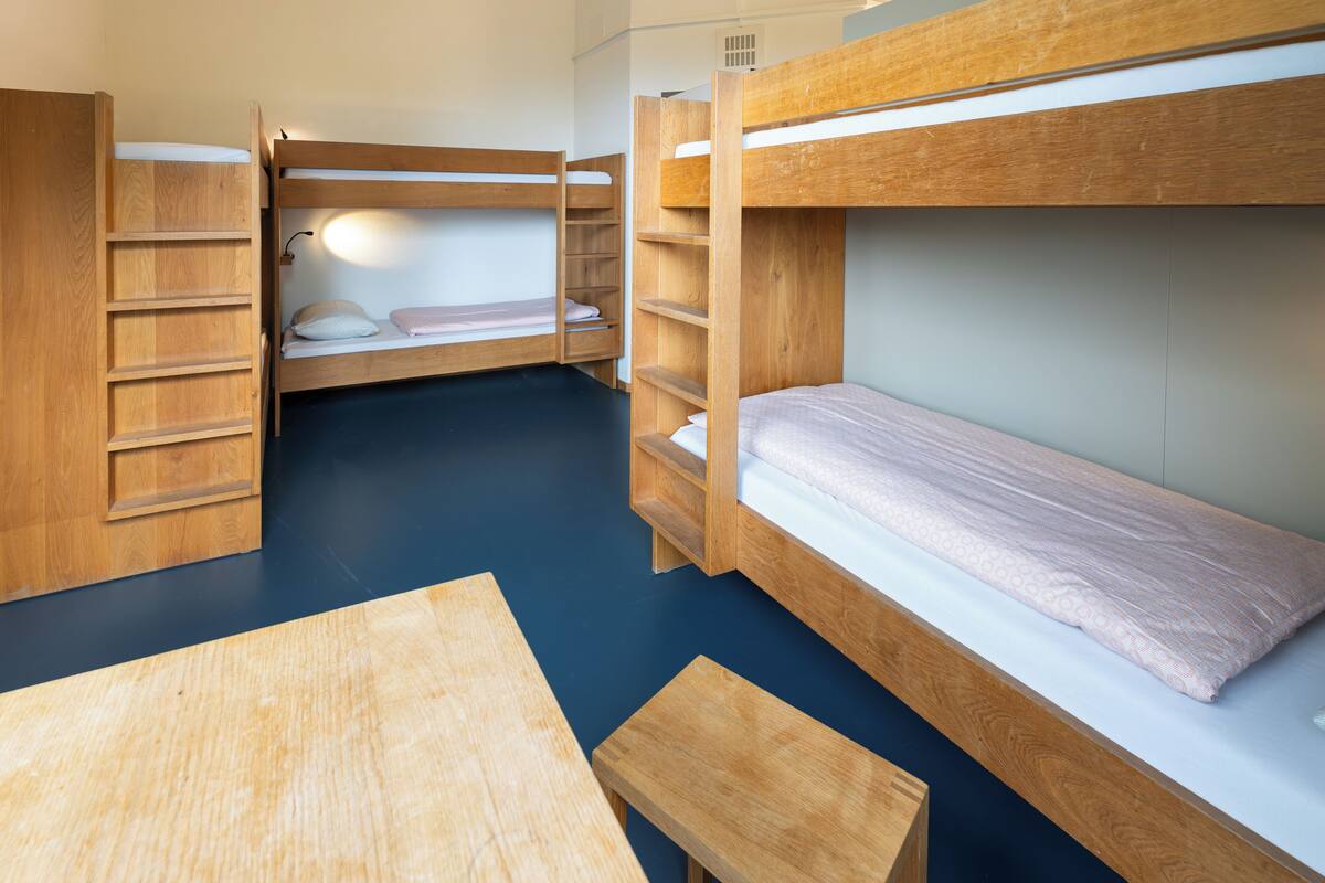 Habitación con literas, una mesa de madera y un banco, estantes empotrados y una lámpara.