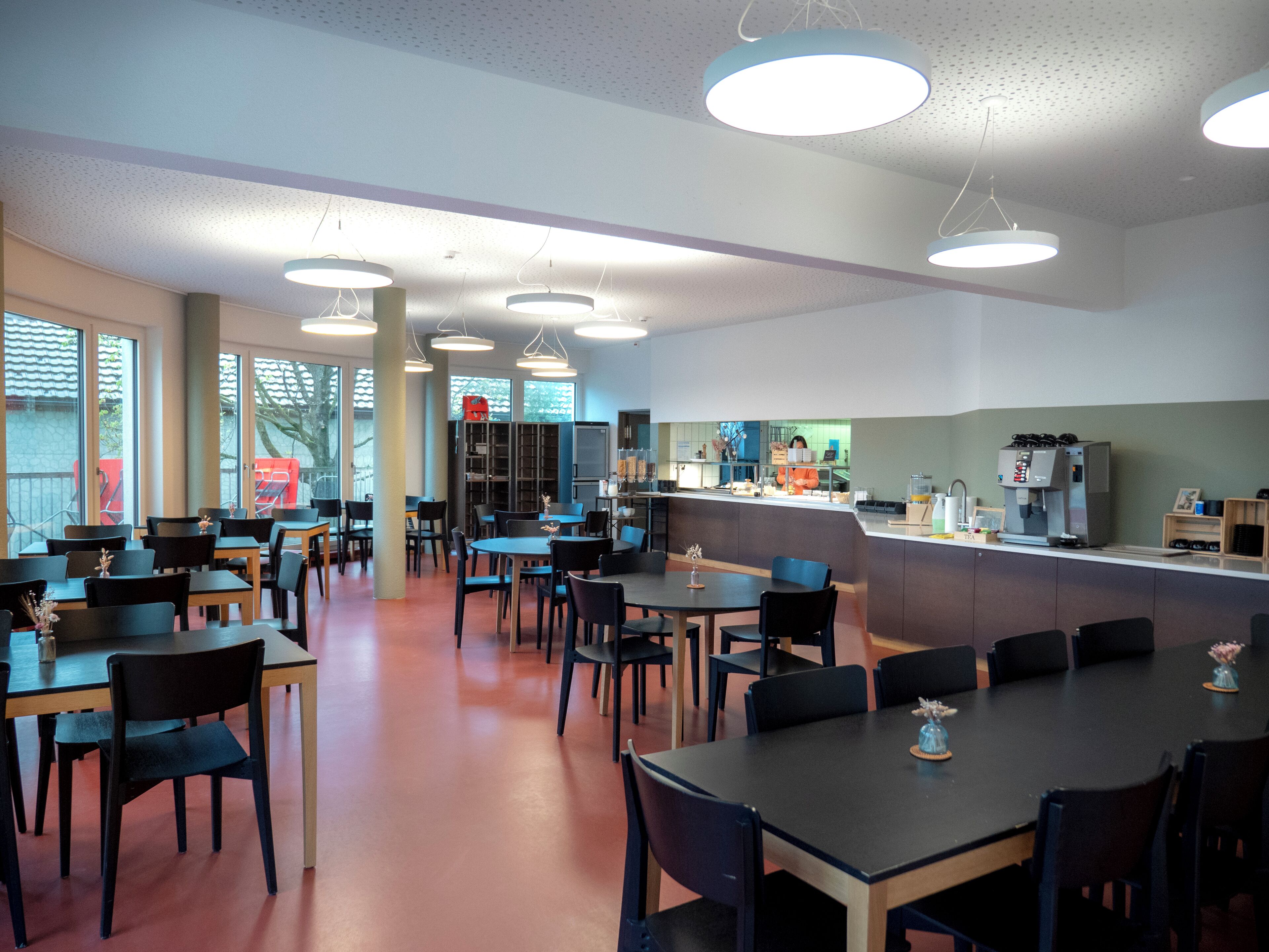 Una amplia cafetería con varias mesas y sillas, un mostrador con máquina de café, y un ventanal que proporciona luz natural.