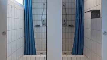 Un baño con espejo, paredes y suelo de azulejos blancos, una ducha con cortina azul y un ventanal que deja entrar luz natural.
