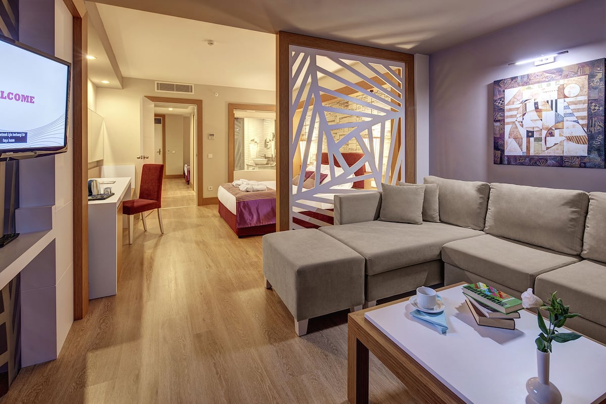 A modern hotel room with a sectional sofa, a desk, a chair, a bed, and a decorative screen.