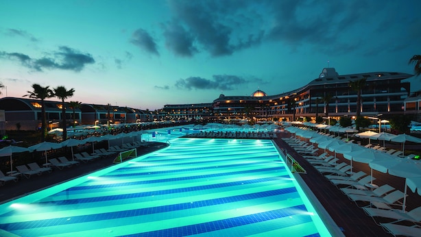 Una amplia piscina interior con suelo de baldosas azules y blancas, rodeada de sillones de descanso y sombrillas, y un hotel al fondo.