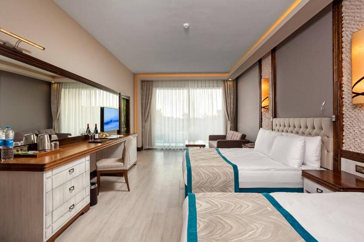 A hotel room with a large bed, a desk with a flat-screen TV, a minibar, and a large window with curtains.