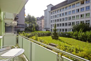 Varanda com mesa e cadeiras de metal, com vista para um jardim bem cuidado e um edifício de vários andares com muitas janelas.