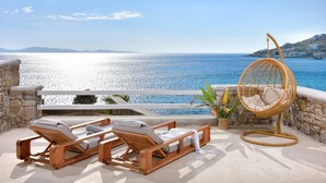 A balcony with wooden lounge chairs and a wicker hanging chair overlooking a coastal view. - Anax Resort & Spa (Mykonos)