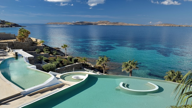 A pool with a curved design overlooking a clear blue sea, surrounded by stone structures and palm trees. - Anax Resort & Spa (Mykonos)