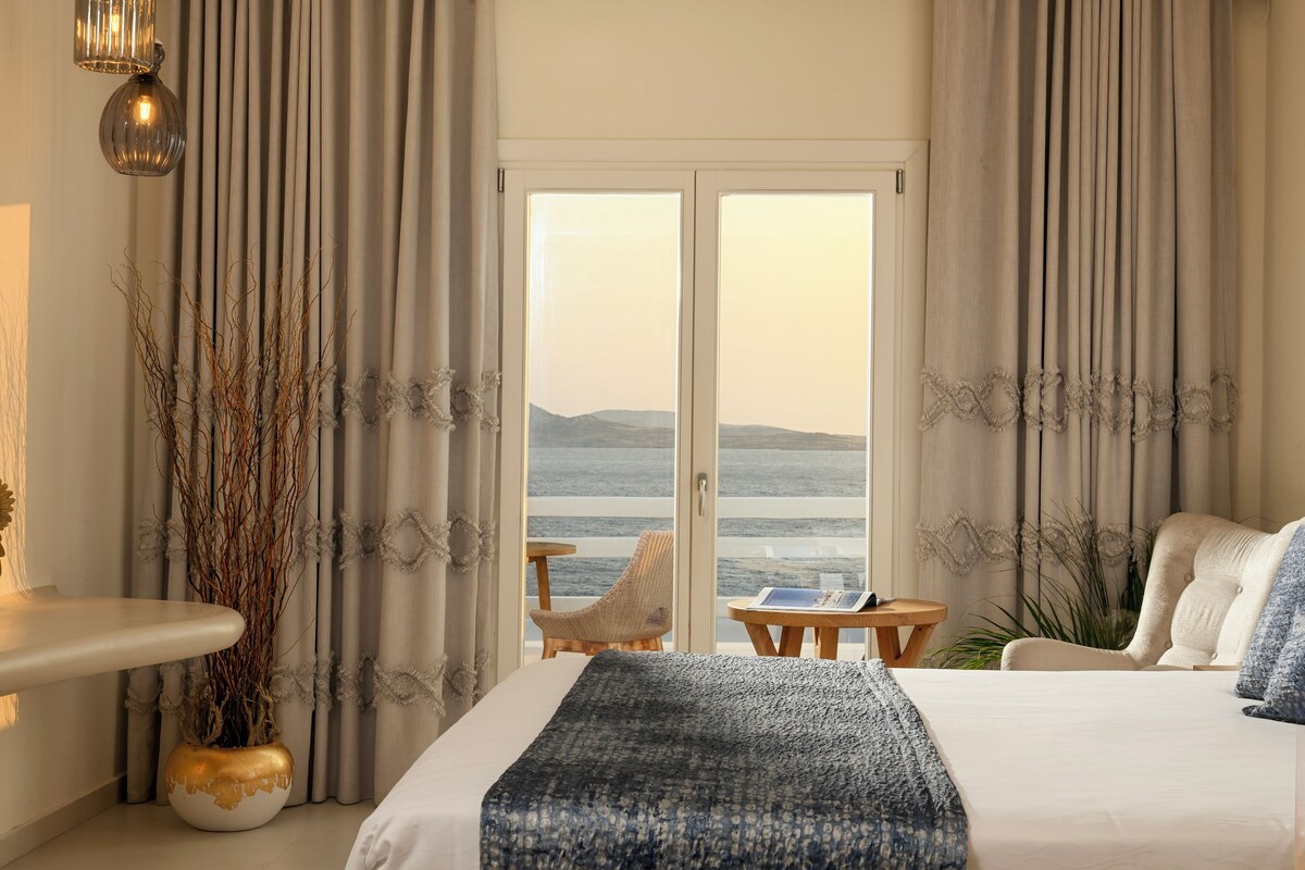 A bedroom with a bed, a nightstand, a window with curtains, a view of the sea, and a potted plant.