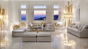 A modern living room with a large sofa, a coffee table, and two side tables. There are three large windows with white curtains, and a view of the sea is visible through them. - Anax Resort & Spa (Mykonos)