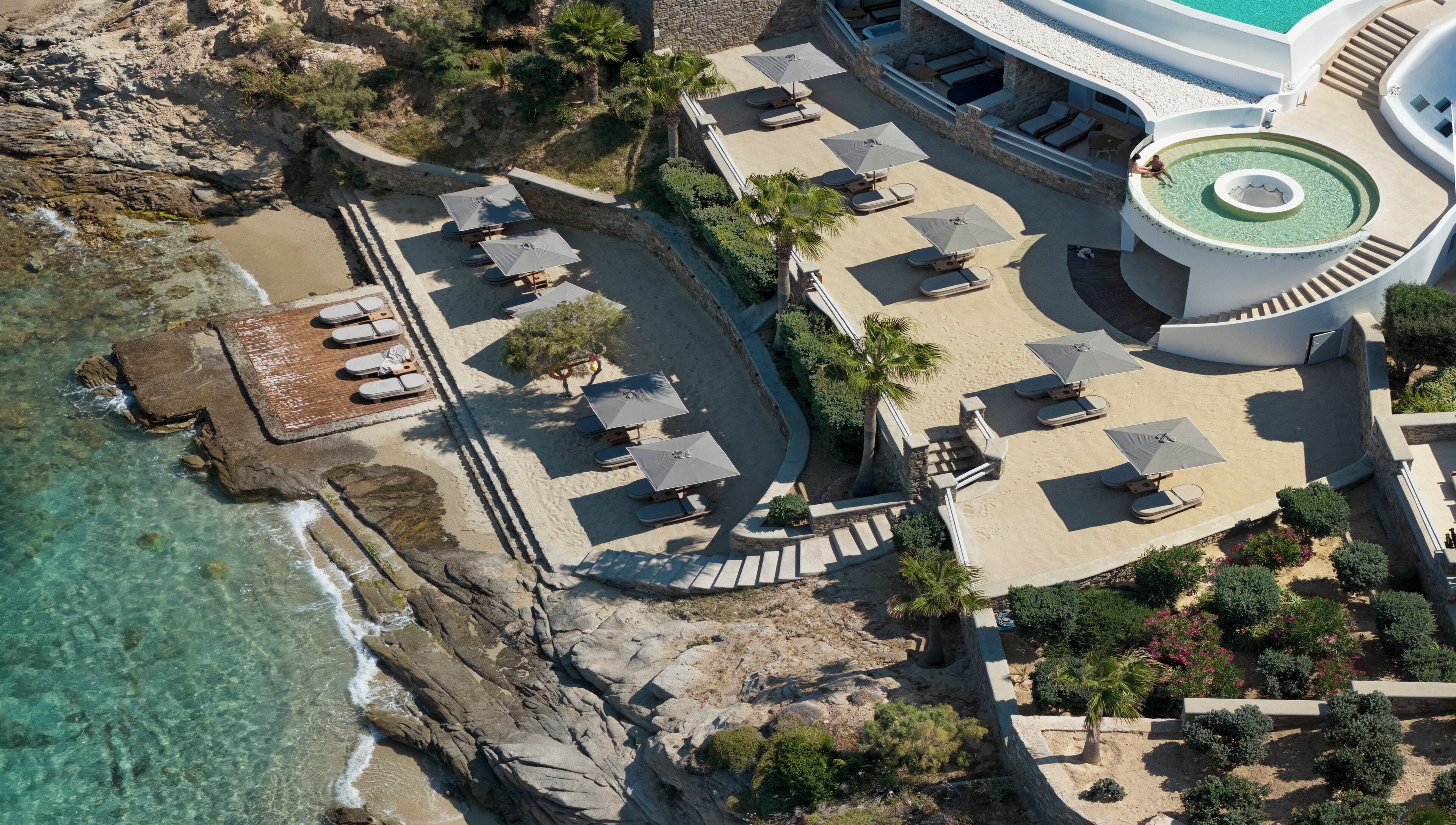 An aerial view of a resort with a beach area, lounge chairs, umbrellas, and a pool.
