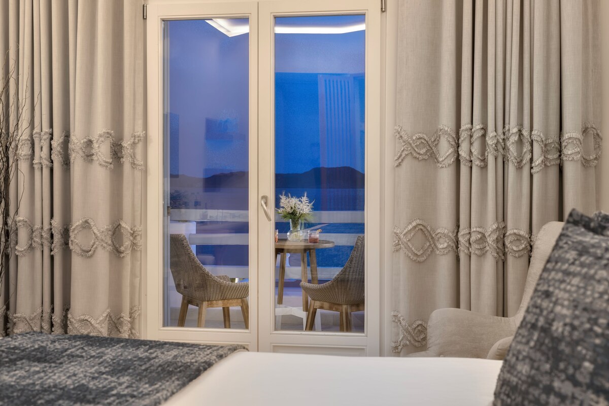 A hotel room with a balcony, two wicker chairs, a small round table with a vase, and curtains with a textured pattern.