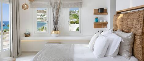 A bedroom with a bed, bedside tables, a window with a view of the sea, and a shelf with decorative items.