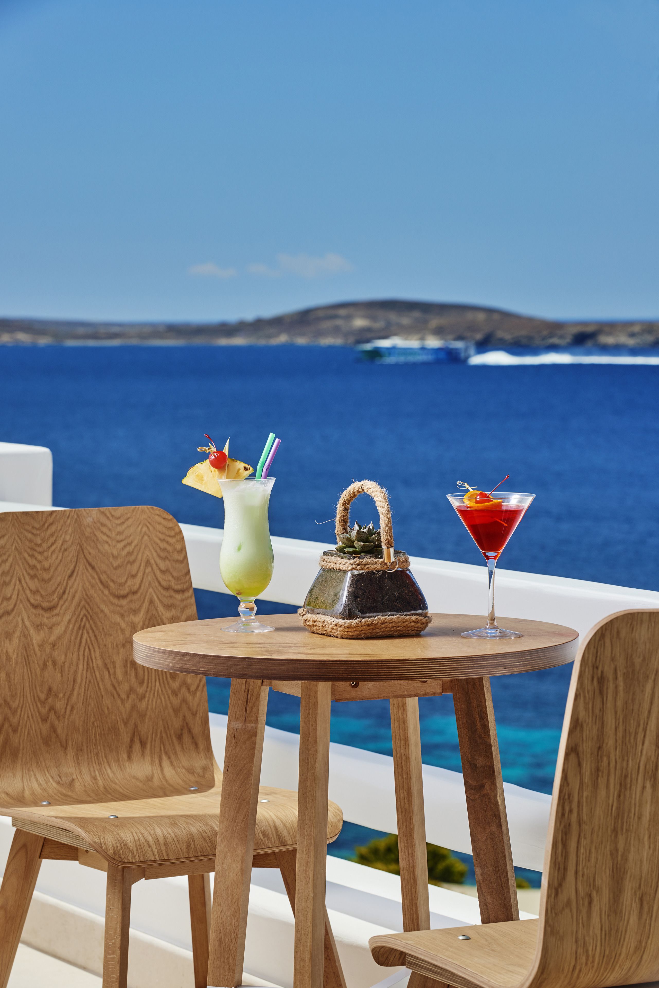 A table with two drinks and a basket on a balcony overlooking the sea.